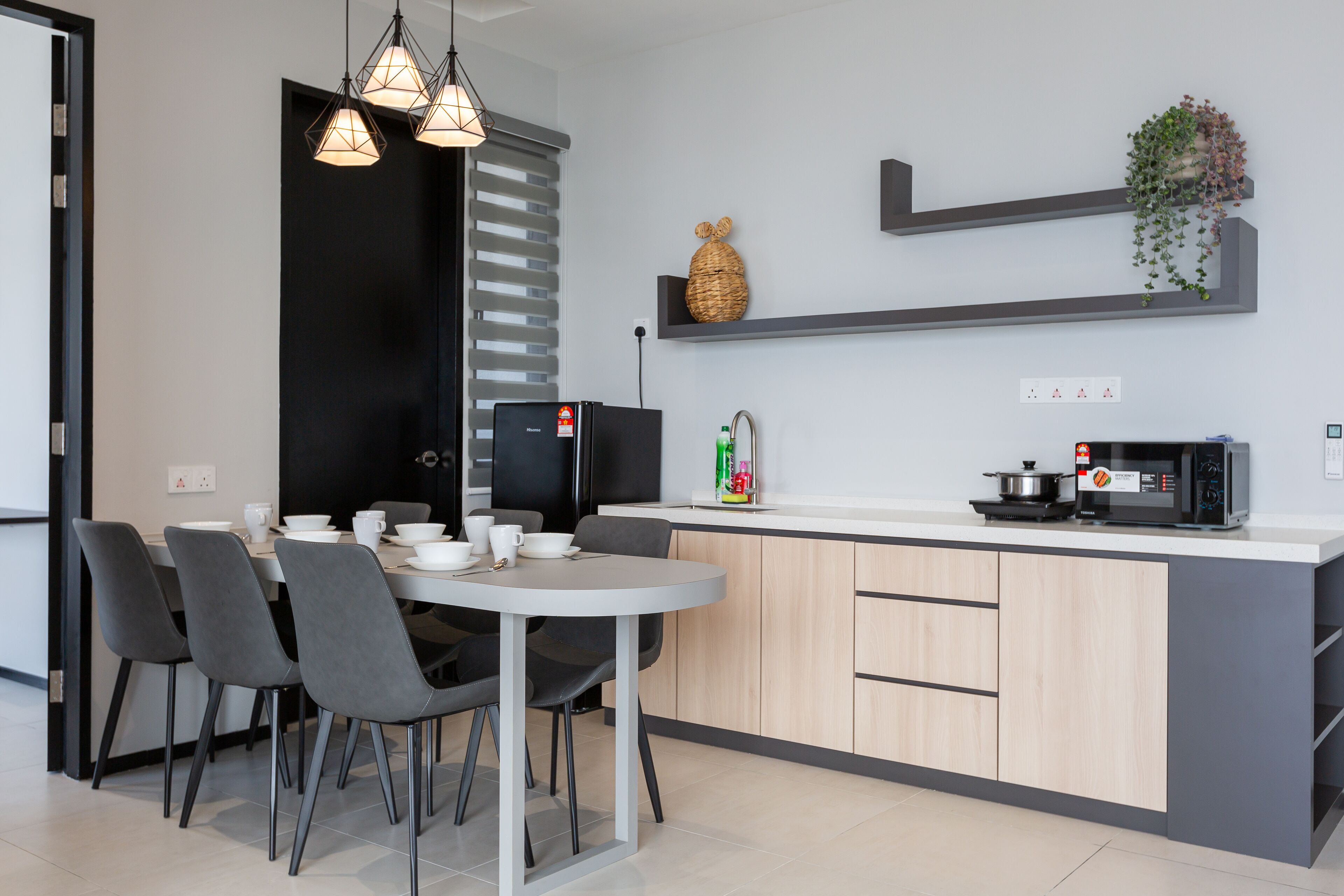 A modern kitchen with a central island, grey chairs, pendant lights, a black refrigerator, a microwave, and a shelf with plants.