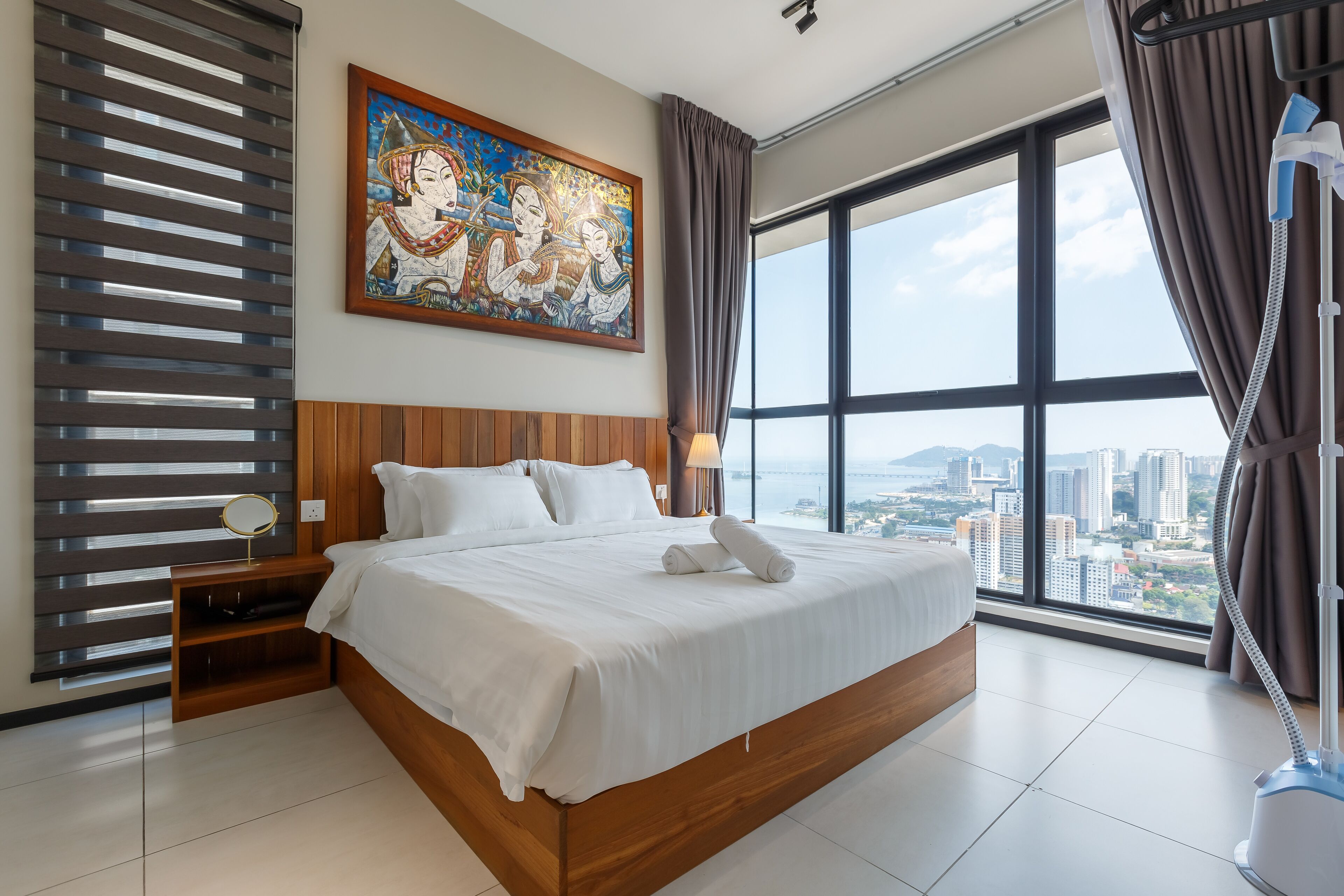 A hotel room with a large bed, a nightstand, a mirror, a painting, and a view of the city through large windows.