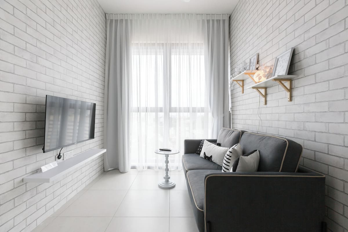 A modern living room with a grey sofa, a flat-screen TV mounted on a white brick wall, and a small white side table.