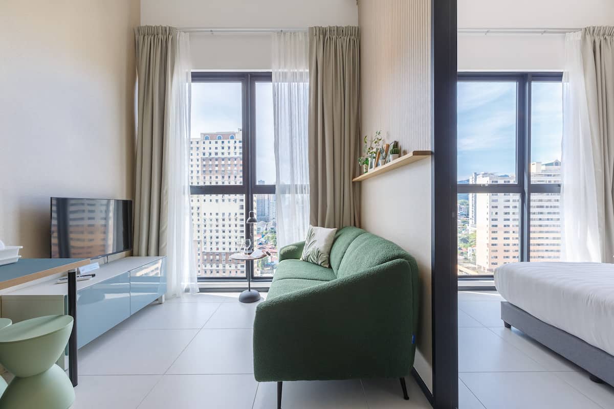 A modern hotel room with a green sofa, a bed, a TV, and a balcony with city views.