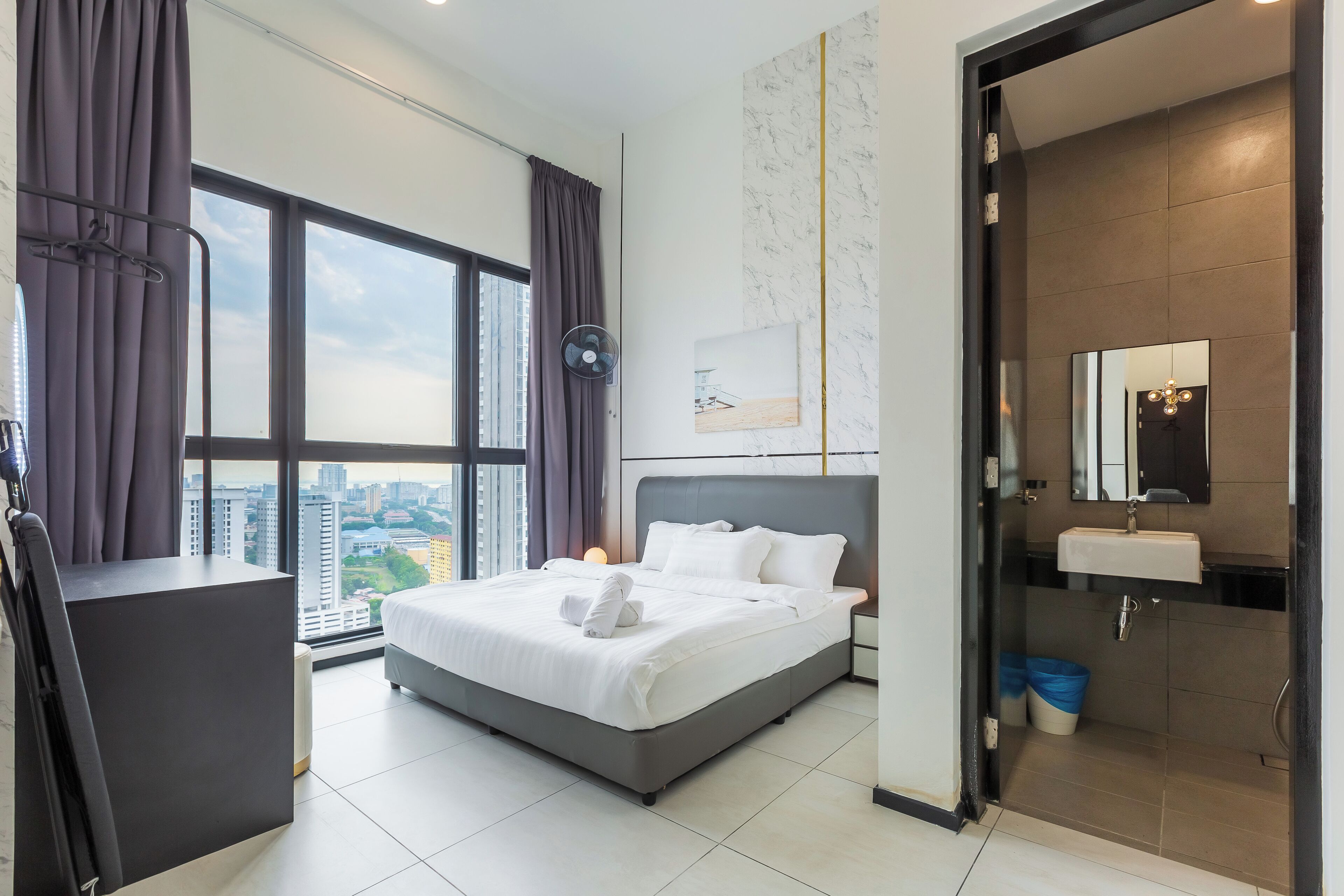 A modern hotel room with a large bed, a desk, a view of the city, and a bathroom with a sink and mirror.