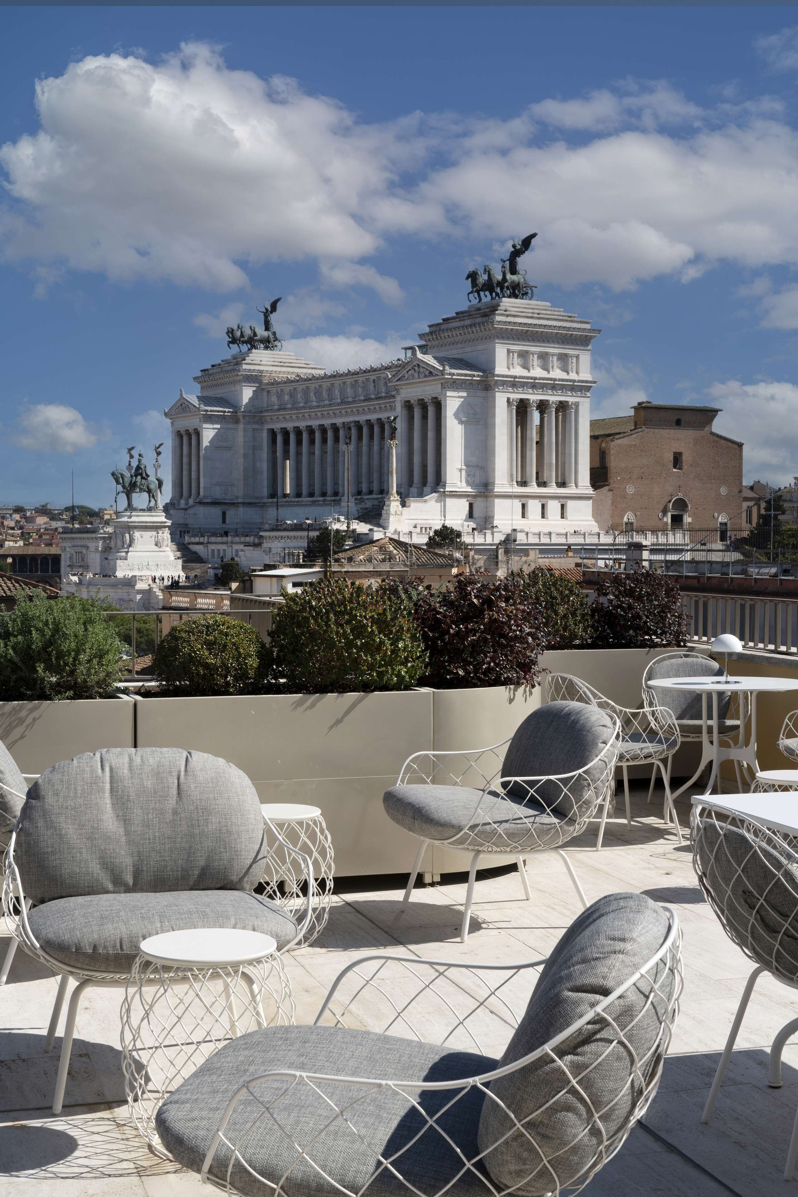 Eine Dachterrasse mit Sitzgelegenheiten im Freien und Blick auf ein klassizistisches Gebäude mit Statuen.