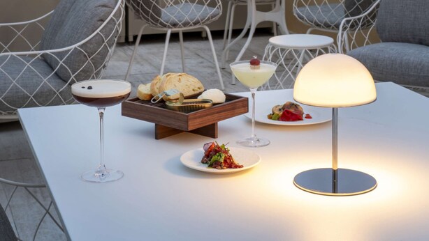 A rooftop terrace with a table set for dining, featuring a lamp, wine glass, and plates of food. In the background, there is a large classical building with statues on top. - Radisson Collection Hotel, Roma Antica (Rome)