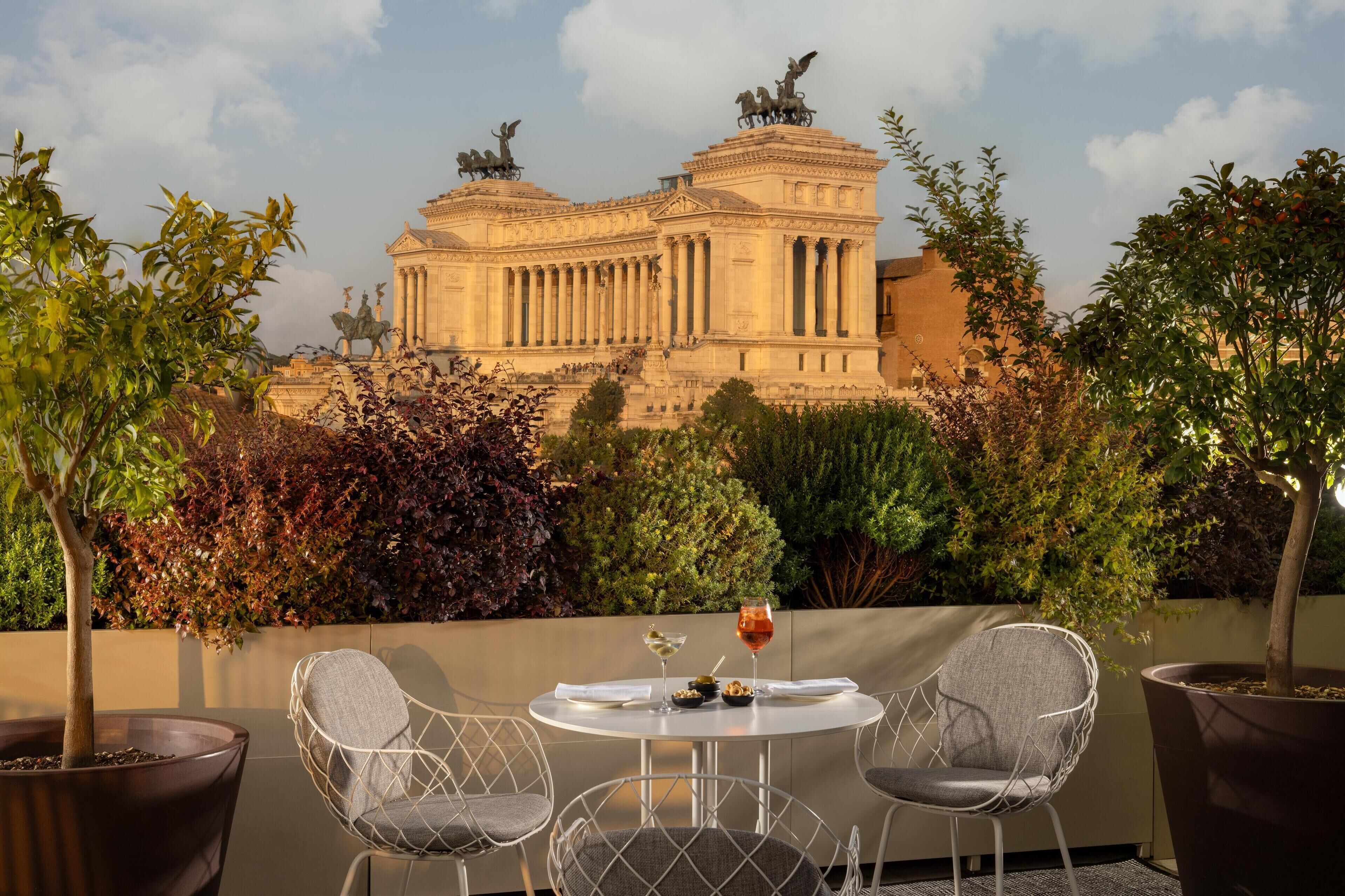 Um terraço com mesa e cadeiras, vista de um edifício clássico com estátuas e diversas plantas.