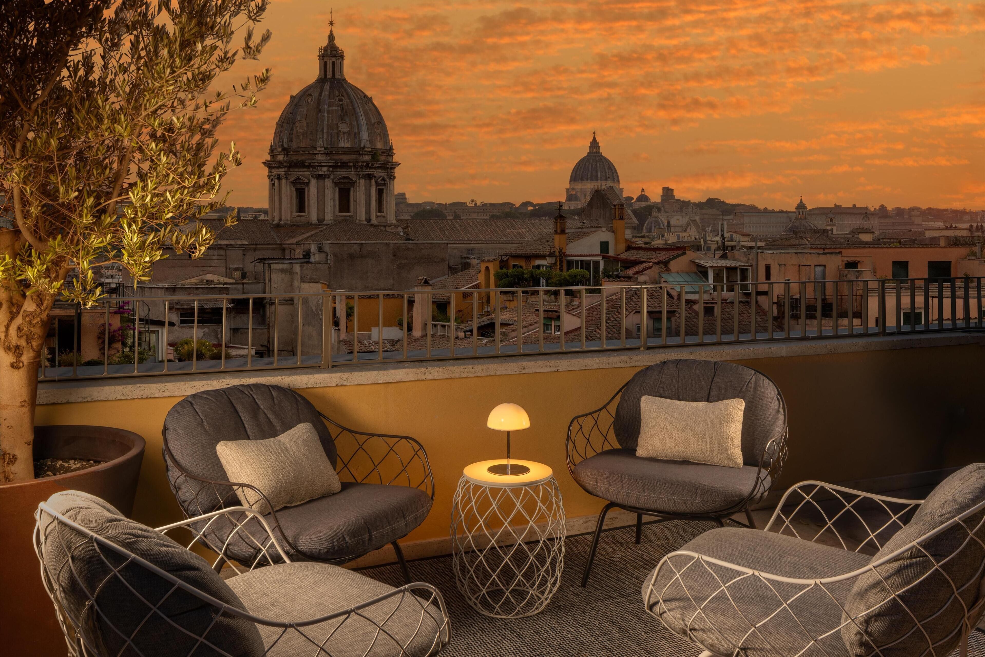 Una terrazza con quattro poltrone imbottite, un tavolino con una lampada e vista su edifici storici e una cupola.