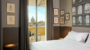 A hotel room with a bed, a chair, a small table, and a view of a cityscape through a large window. - Radisson Collection Hotel, Roma Antica (Rome)