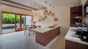 A modern kitchen with a large island, pendant lights, and a view of a lush garden through glass doors.