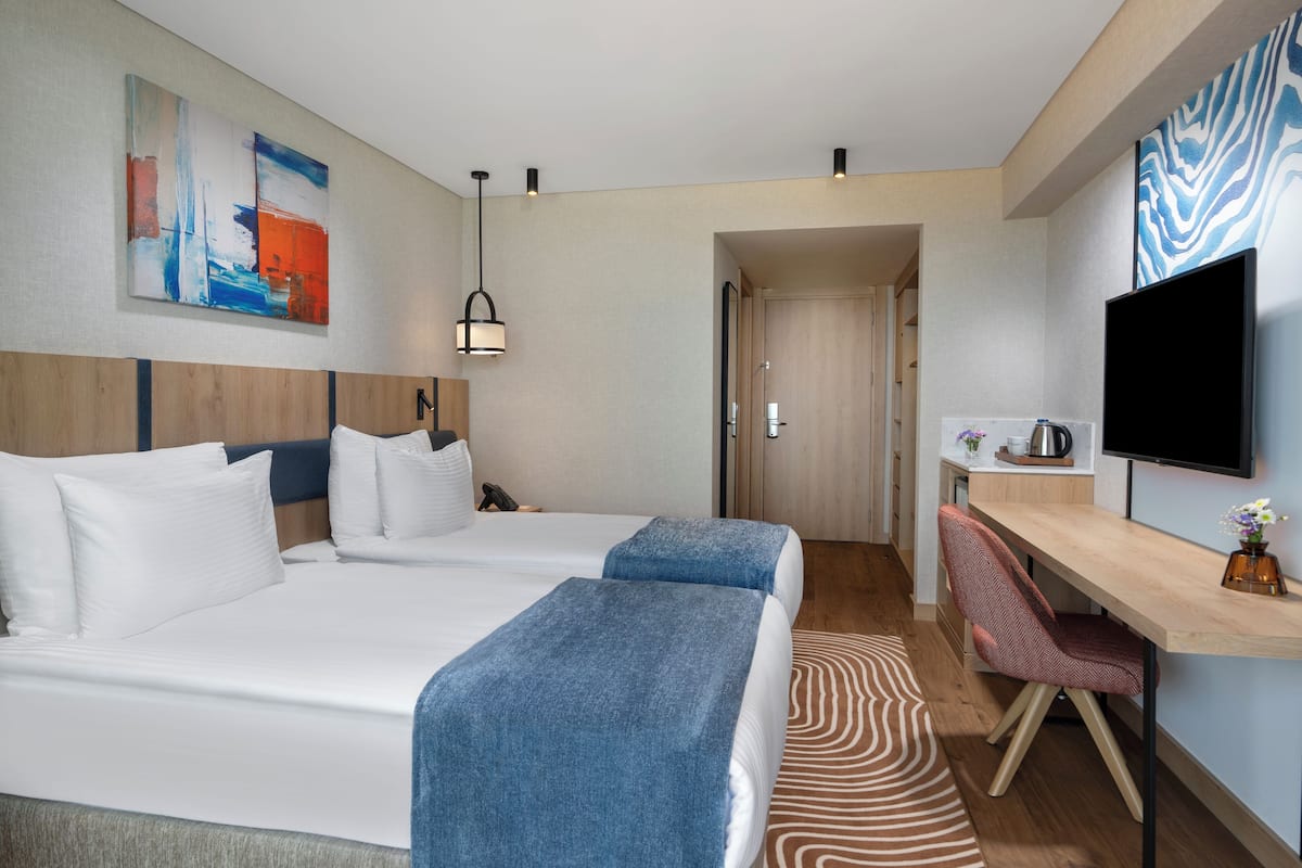 A hotel room with a bed, a desk, a chair, a TV, and a door leading to another room.