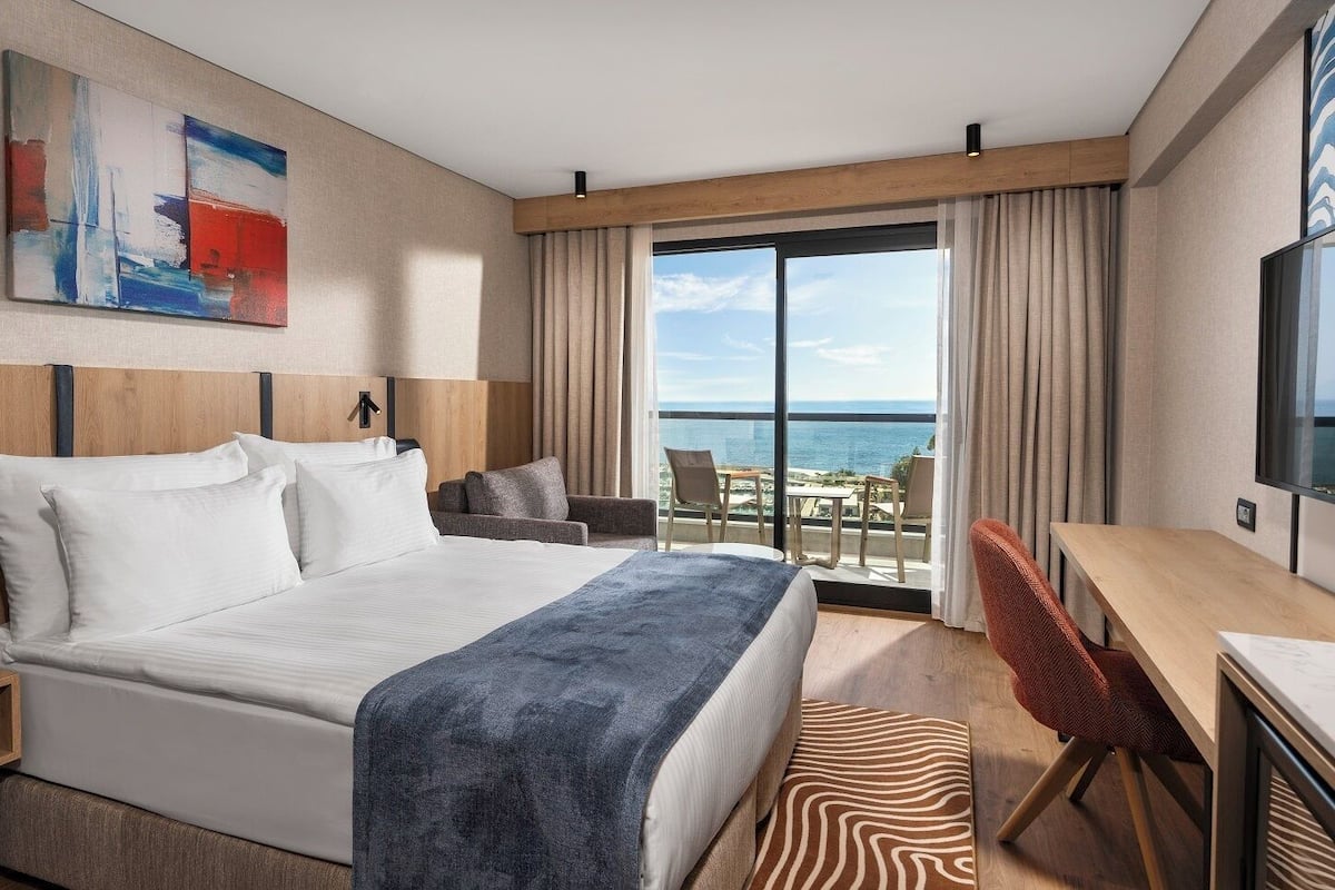 A hotel room with a large bed, a desk, a chair, and a balcony with a view of the sea.