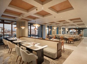 A modern dining area with high ceilings, large windows, and a variety of seating arrangements including booths and tables. - Elite World Kuşadası (Kusadasi)