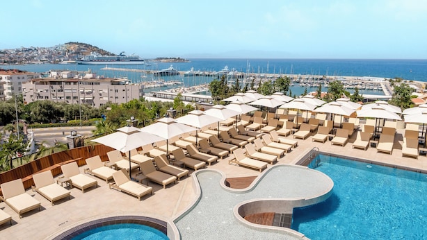 Zona de piscina con sillones de descanso y sombrillas, cielo despejado y vista al mar y edificios al fondo.