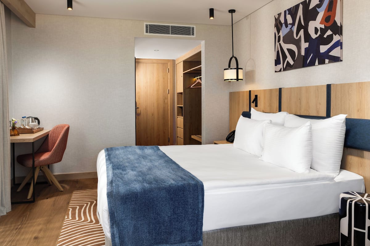 A hotel room with a bed, a desk, a chair, a wardrobe, and a painting on the wall.