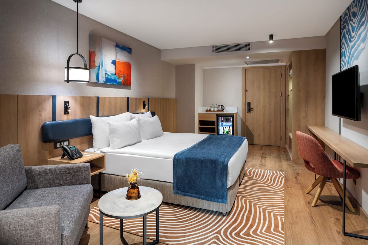 A modern hotel room with a large bed, a desk, a chair, a small table, and a TV.