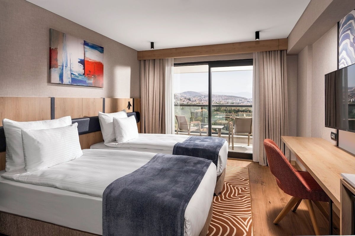 A hotel room with two beds, a desk, a chair, a TV, and a large window with a city view.