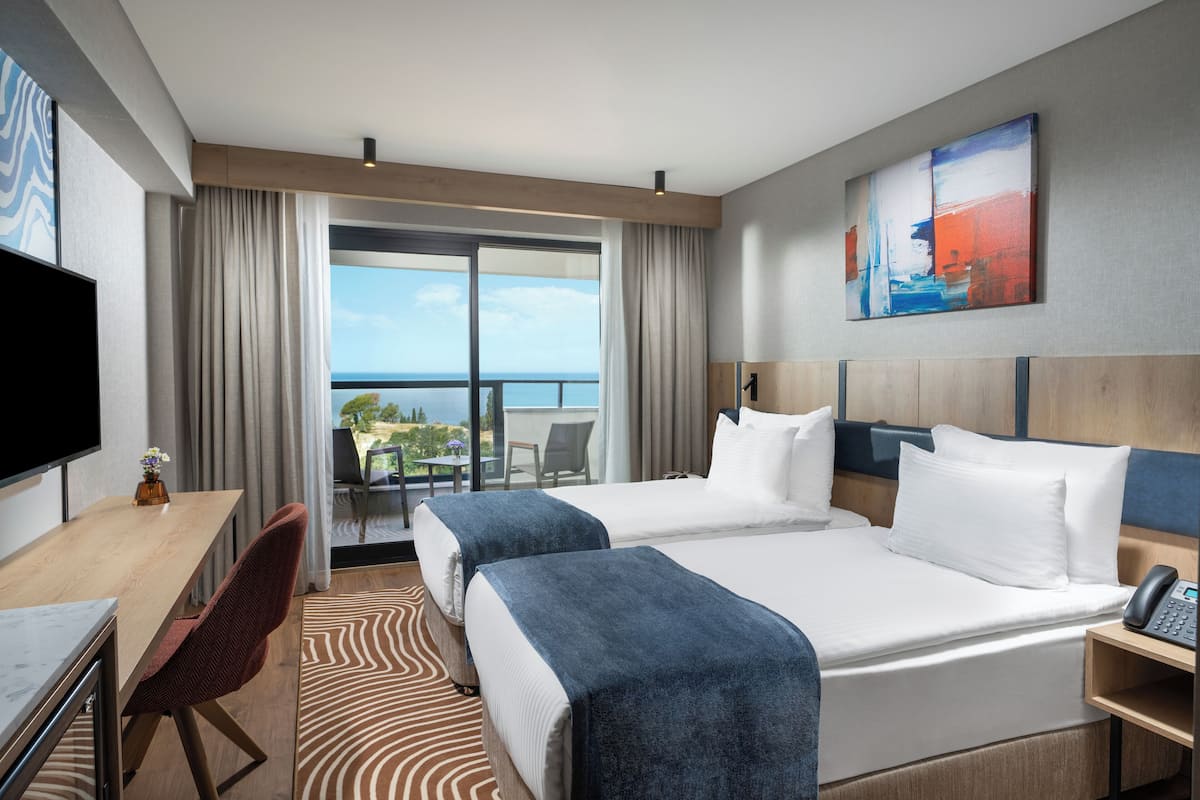 A hotel room with two beds, a desk, a chair, a TV, and a balcony with a view of the sea.