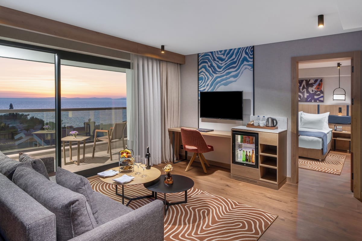 A modern hotel room with a large window, a flat-screen TV, a desk with a chair, a sofa, and a minibar.