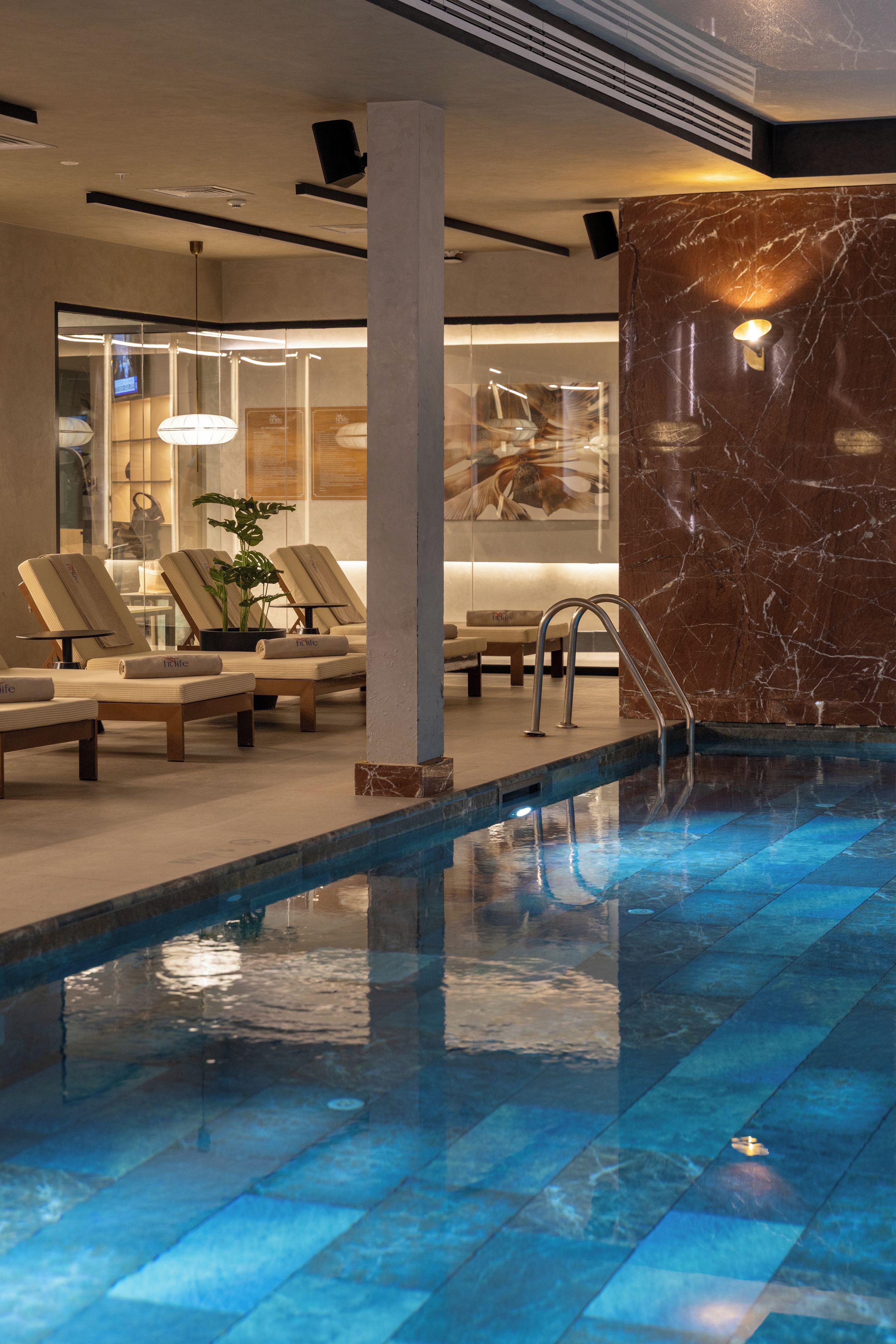 Zona de piscina con sillas de descanso, pared de mármol y un separador de vidrio.