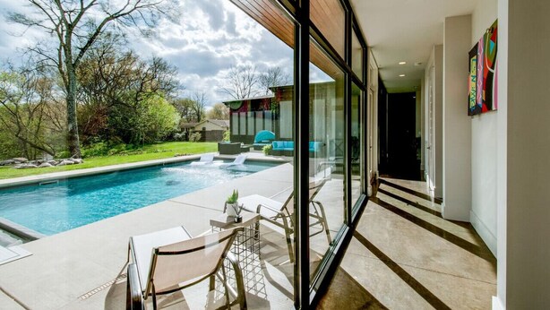 A modern pool area with clear glass walls, a view of the backyard, and a well-maintained lawn. - Unique Modern Ranch with Pool, Hot Tub, Grill & Fireplace (Nashville)