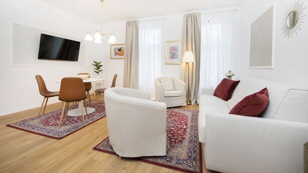 A modern living room with a white sofa, a dining area with a table and chairs, a TV mounted on the wall, and a large window with curtains. - Stylish apartment 2 min from the Dragon Bridge (Ljubljana)