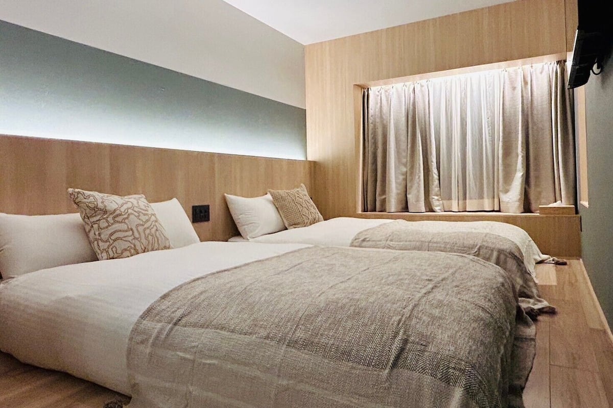 Una habitación de hotel moderna con dos camas, cabecero de madera, ventana grande con cortinas y suelo de color claro.