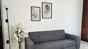 Um sofá cinzento num quarto com parede branca, candeeiro de chão preto e duas obras de arte com folhas emolduradas.