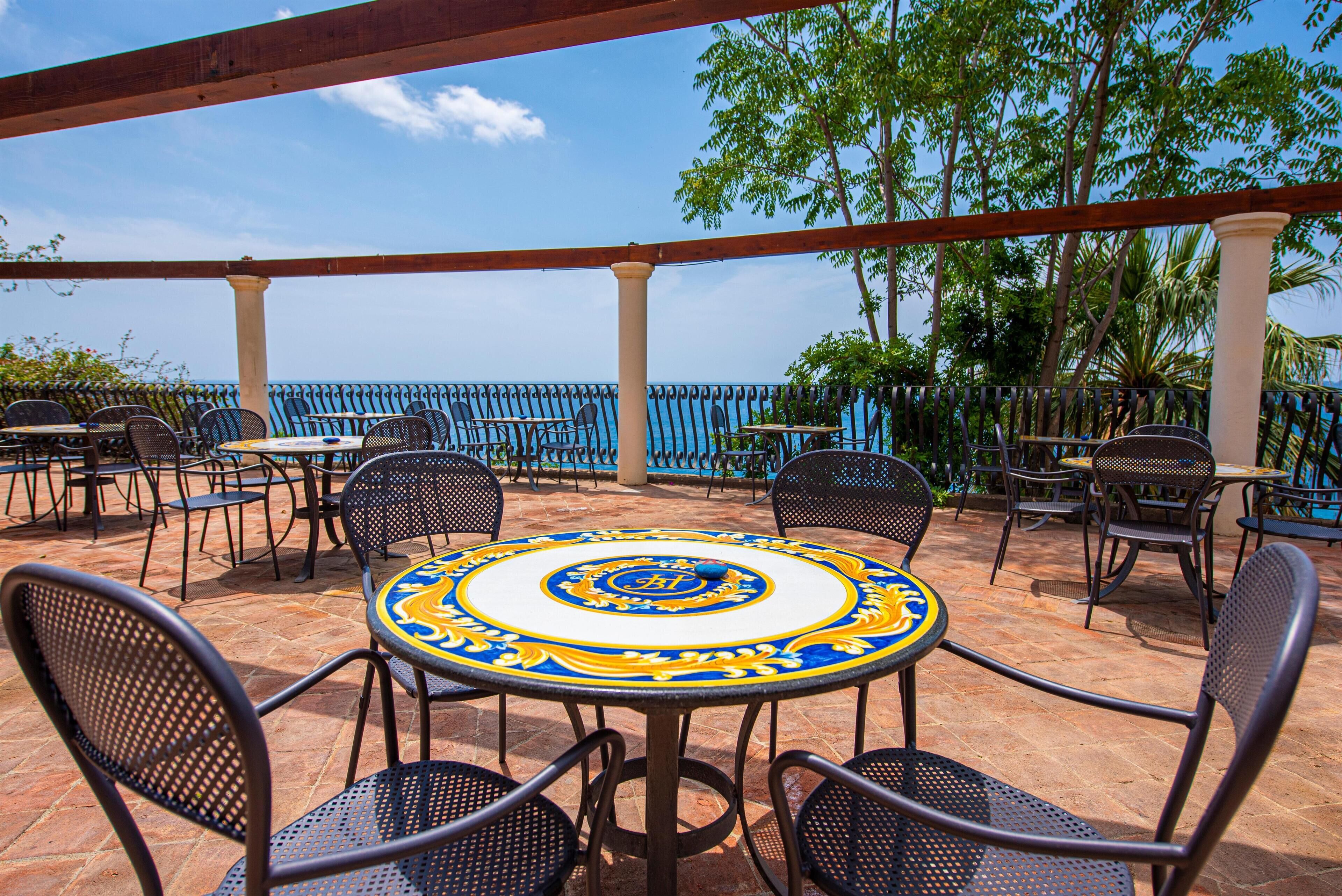 Un patio al aire libre con mesas y sillas redondas, vista al mar y barandal azul.