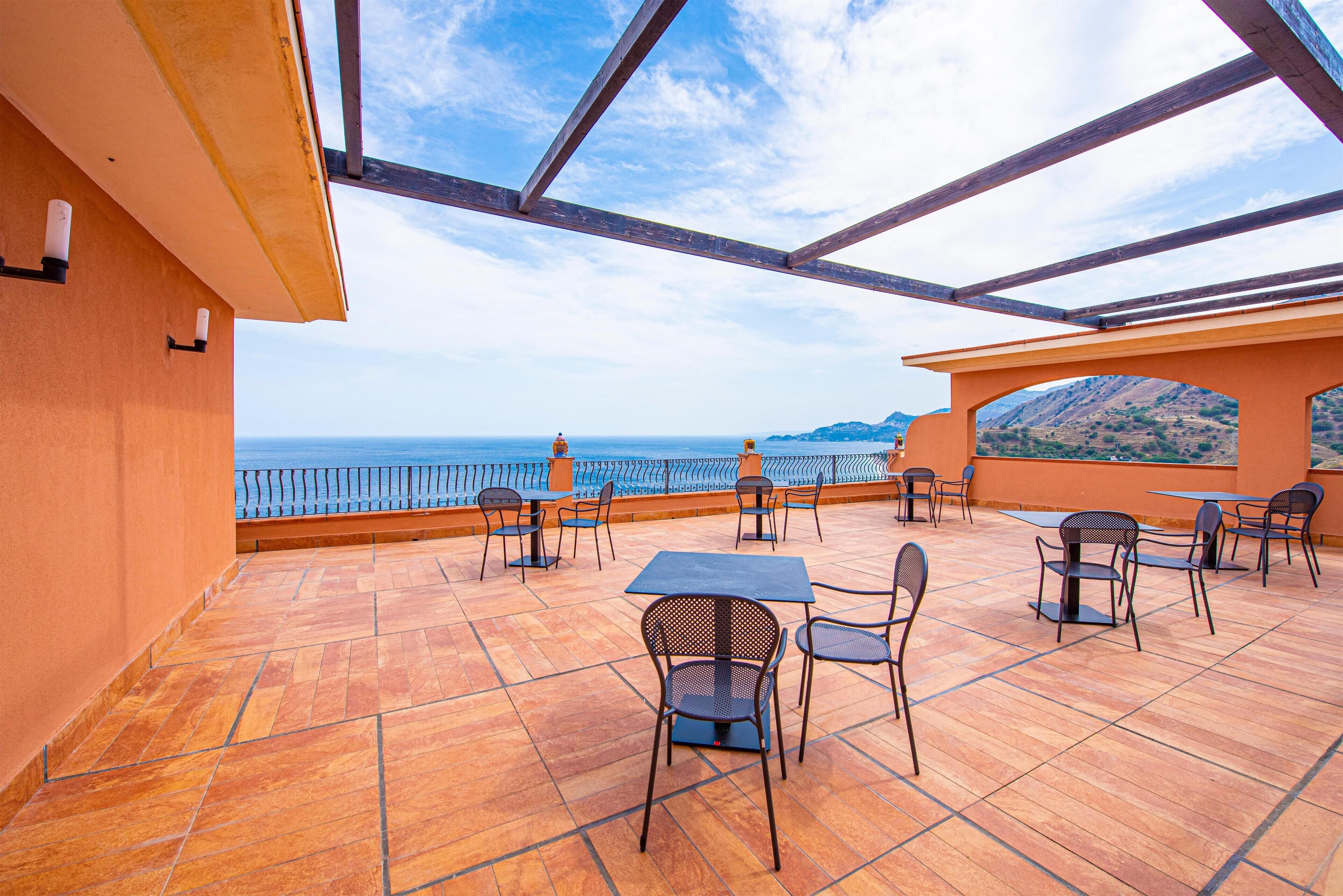 An outdoor terrace with a view of the sea, featuring tables and chairs.