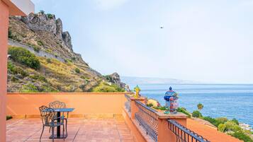 Una terrazza con tavolo e sedie, un parapetto e vista sul mare e su un pendio roccioso.
