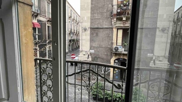 Une vue à travers une fenêtre ornée d’un portail en fer forgé, donnant sur une rue bordée d’immeubles et où est garée une voiture.