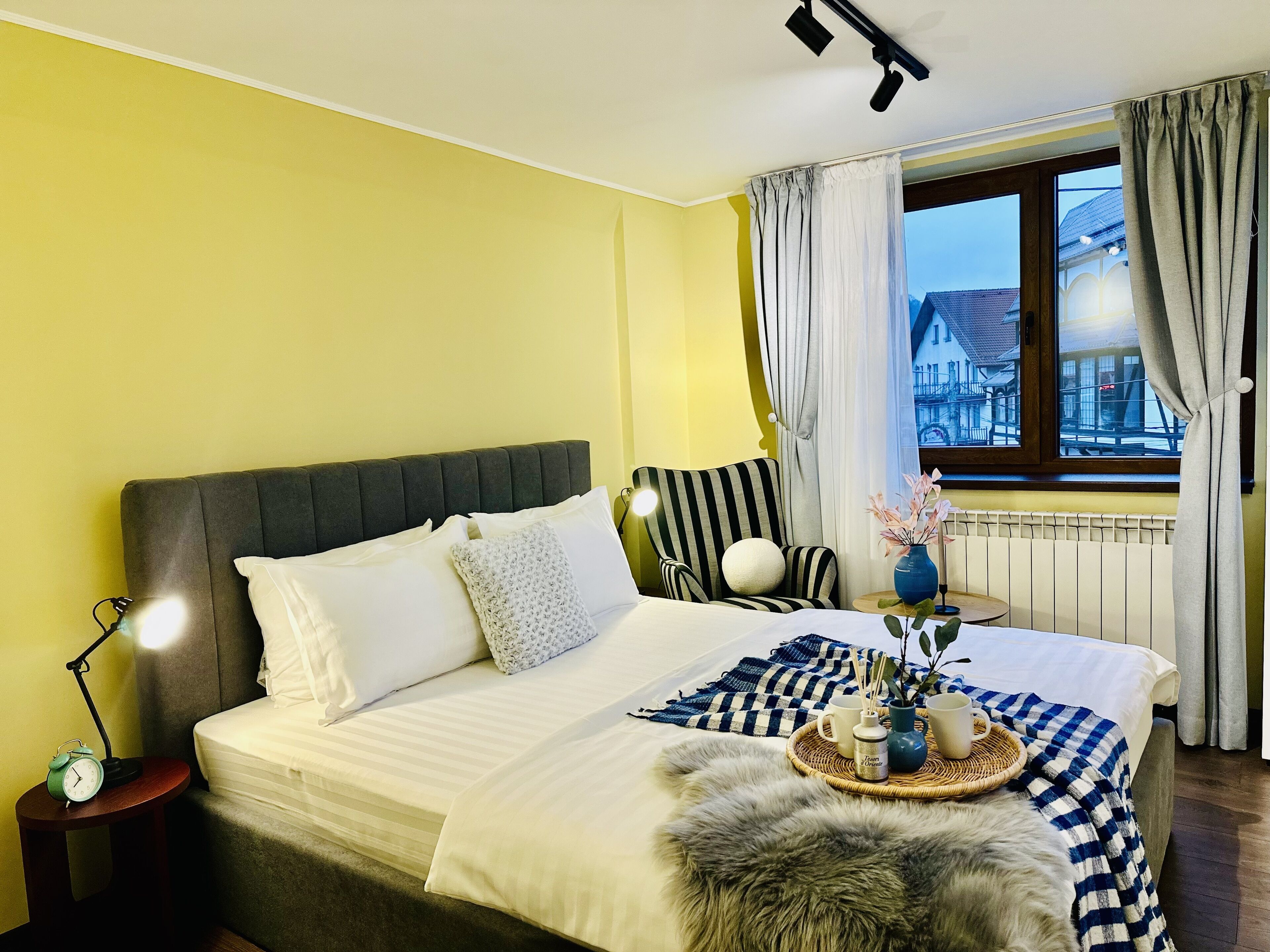A neatly made bed with white linens, a striped armchair, a small table with a tray of items, and a window with curtains.