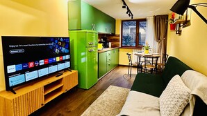 A modern living room with a flat-screen TV, a green refrigerator, a dining area with a table and chairs, and a sofa.