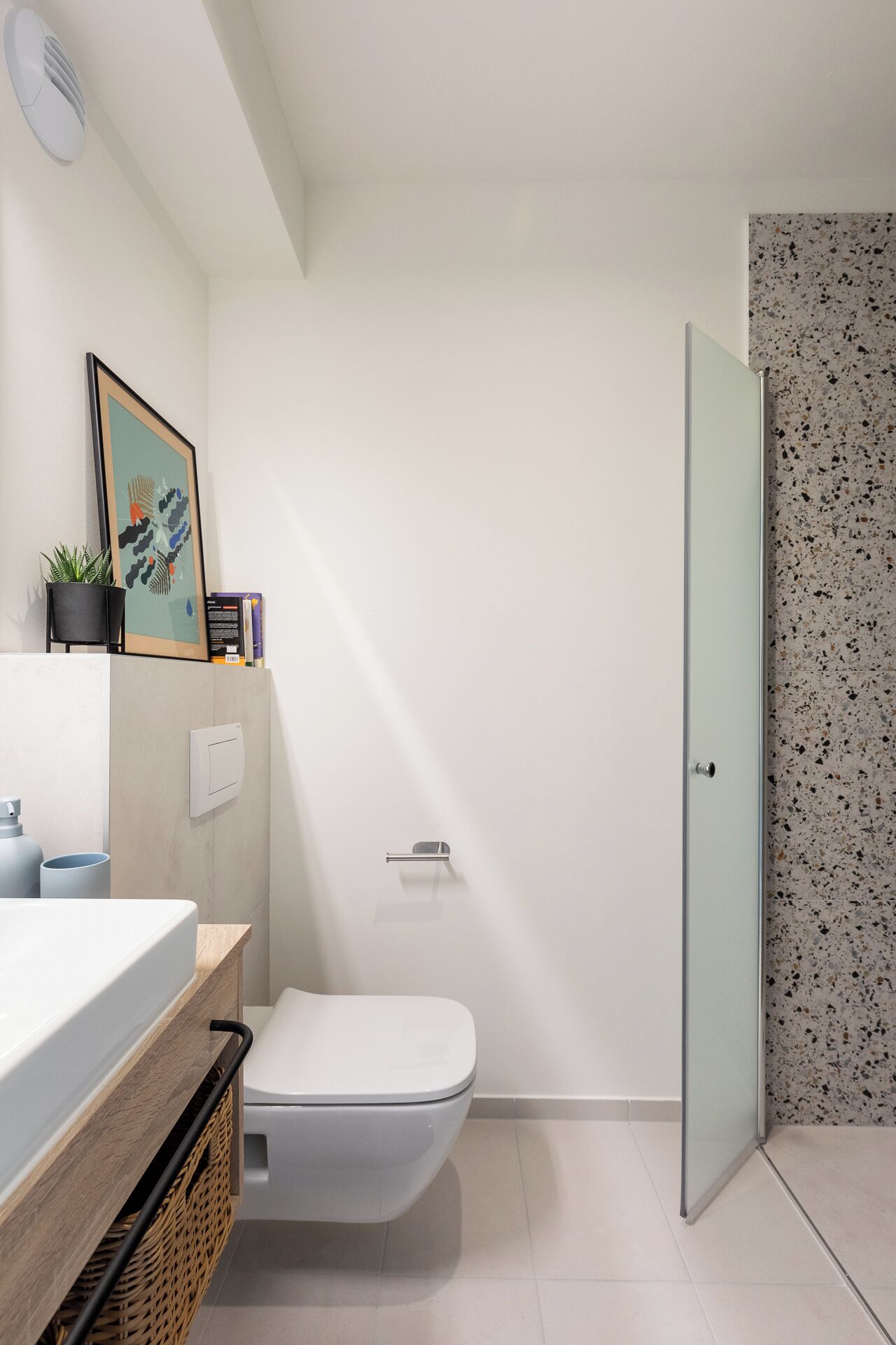 Un baño moderno con inodoro blanco, lavamanos con encimera de madera, una obra de arte enmarcadasobre el lavamanos y una puerta con salpicadero de azulejos veteados.