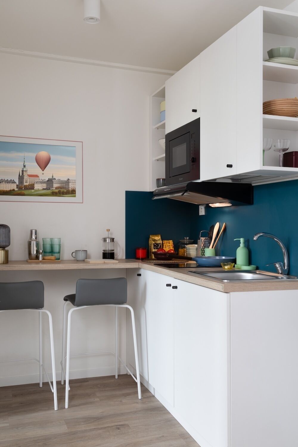 Cocina compacta con armarios blancos, salpicadero verde azulado, microondas y un lavamanos. Hay dos taburetes de bar y un cuadro con una vista urbana en la pared.