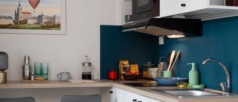 Cocina compacta con armarios blancos, salpicadero verde azulado, microondas y un lavamanos. Hay dos taburetes de bar y un cuadro con una vista urbana en la pared.