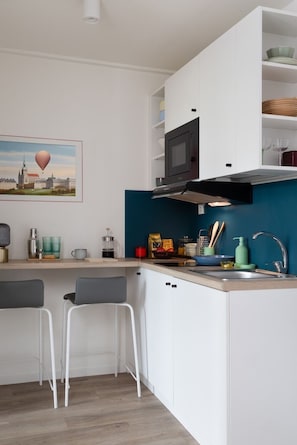 A compact kitchen with white cabinets, a teal backsplash, a microwave, and a sink. There are two bar stools and a painting of a cityscape on the wall. - MILESTONE Krakow Center Student Living by Tribera (Kraków)