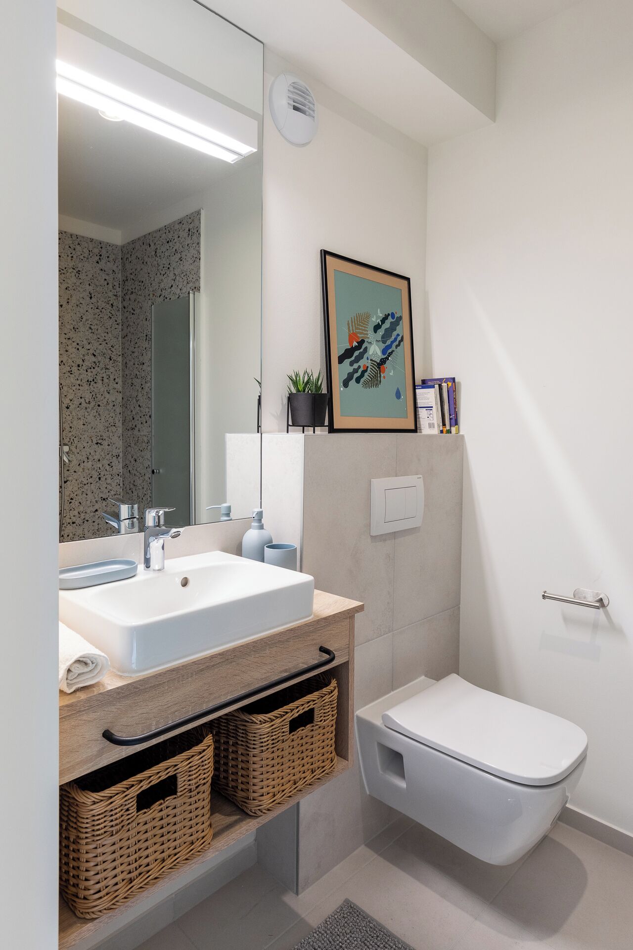 A modern bathroom with a white sink, mirror, and toilet. There are wicker baskets under the sink and a framed picture on the wall.