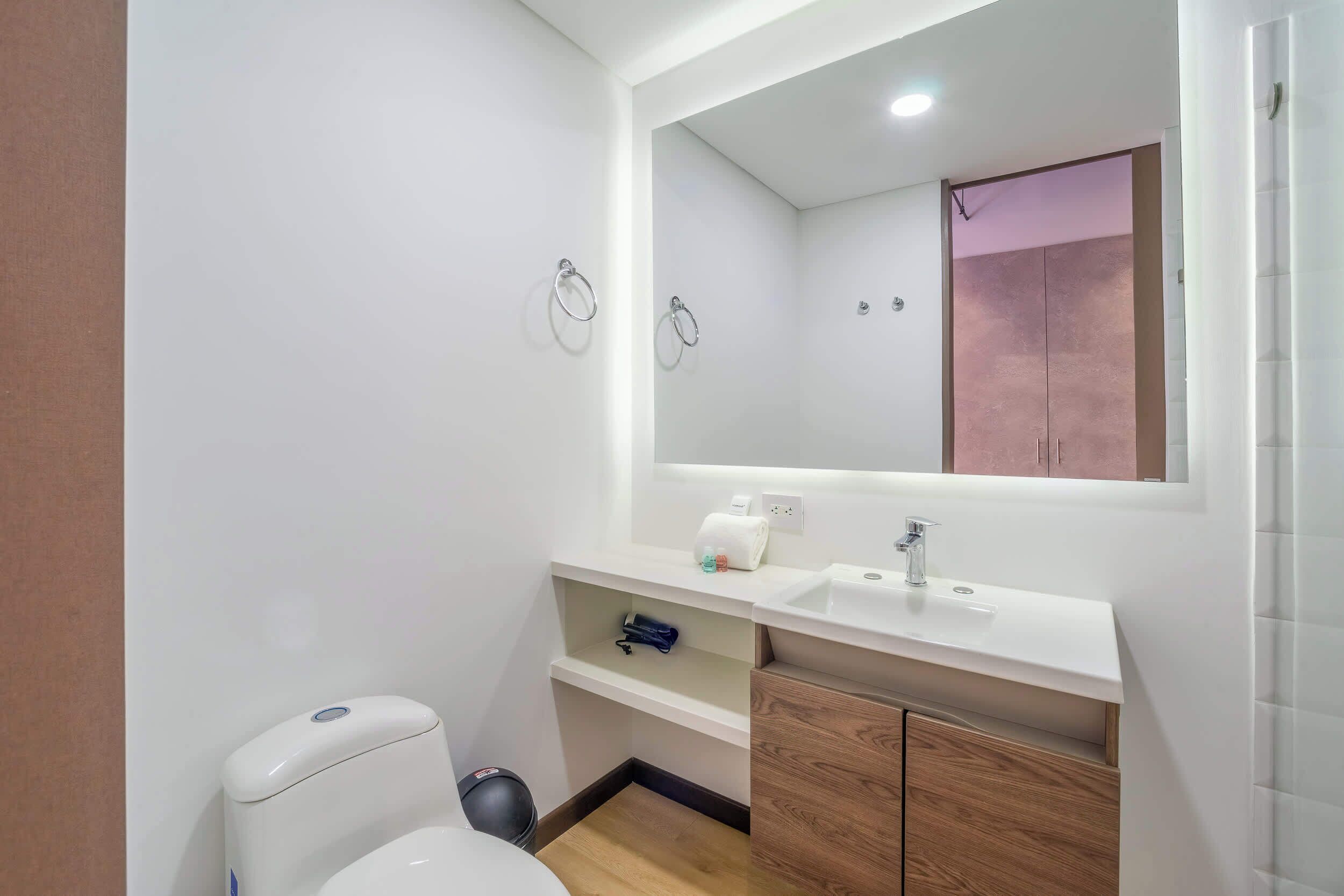 A small, modern bathroom with a white toilet, a sink, and a mirror. There are two towel rings on the wall, and a small shelf under the mirror.