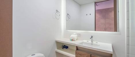 A small, modern bathroom with a white toilet, a sink, and a mirror. There are two towel rings on the wall, and a small shelf under the mirror.