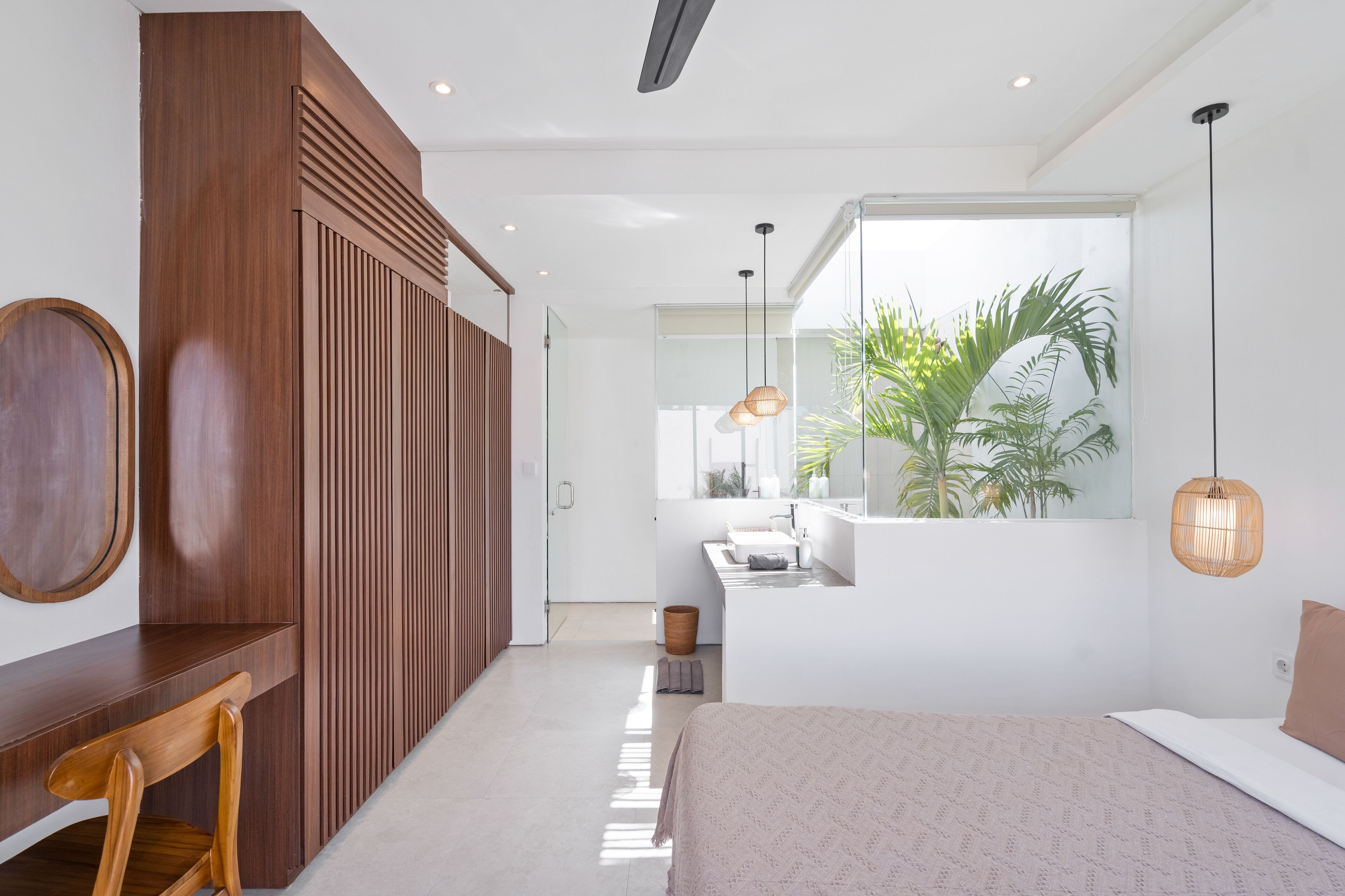 A modern bedroom with a large bed, a wooden desk, a mirror, and a view of a bathroom and plants through a window.