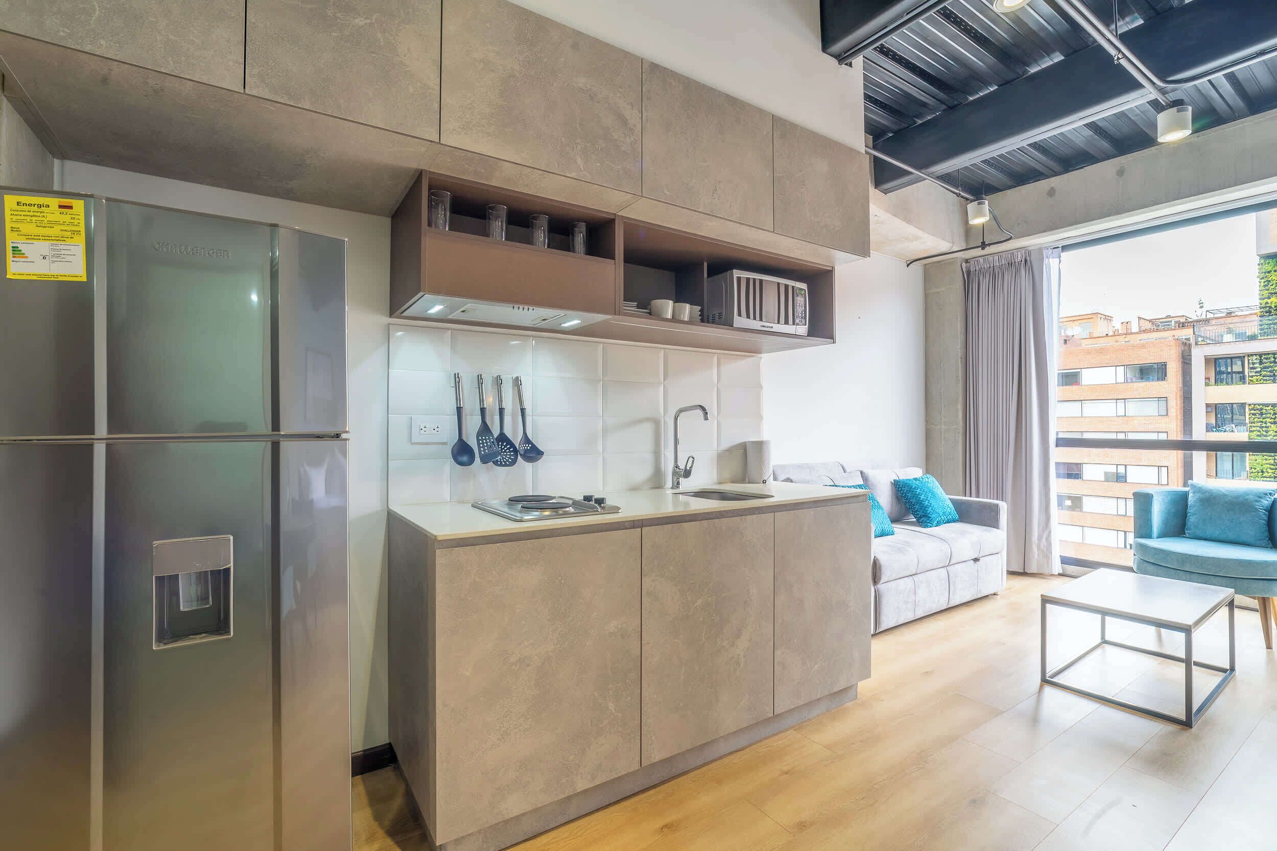 A modern kitchen with a built-in refrigerator, a sink, and a countertop with utensils. There is a sofa and a coffee table in the adjacent living area.