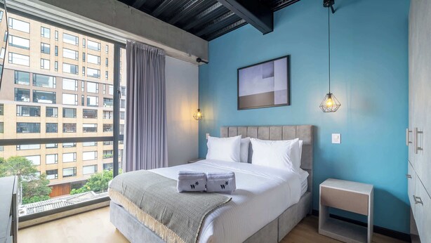 Habitació d'hotel moderna amb un llit gran, vista a un paisatge urbà i una petita taula de nit.