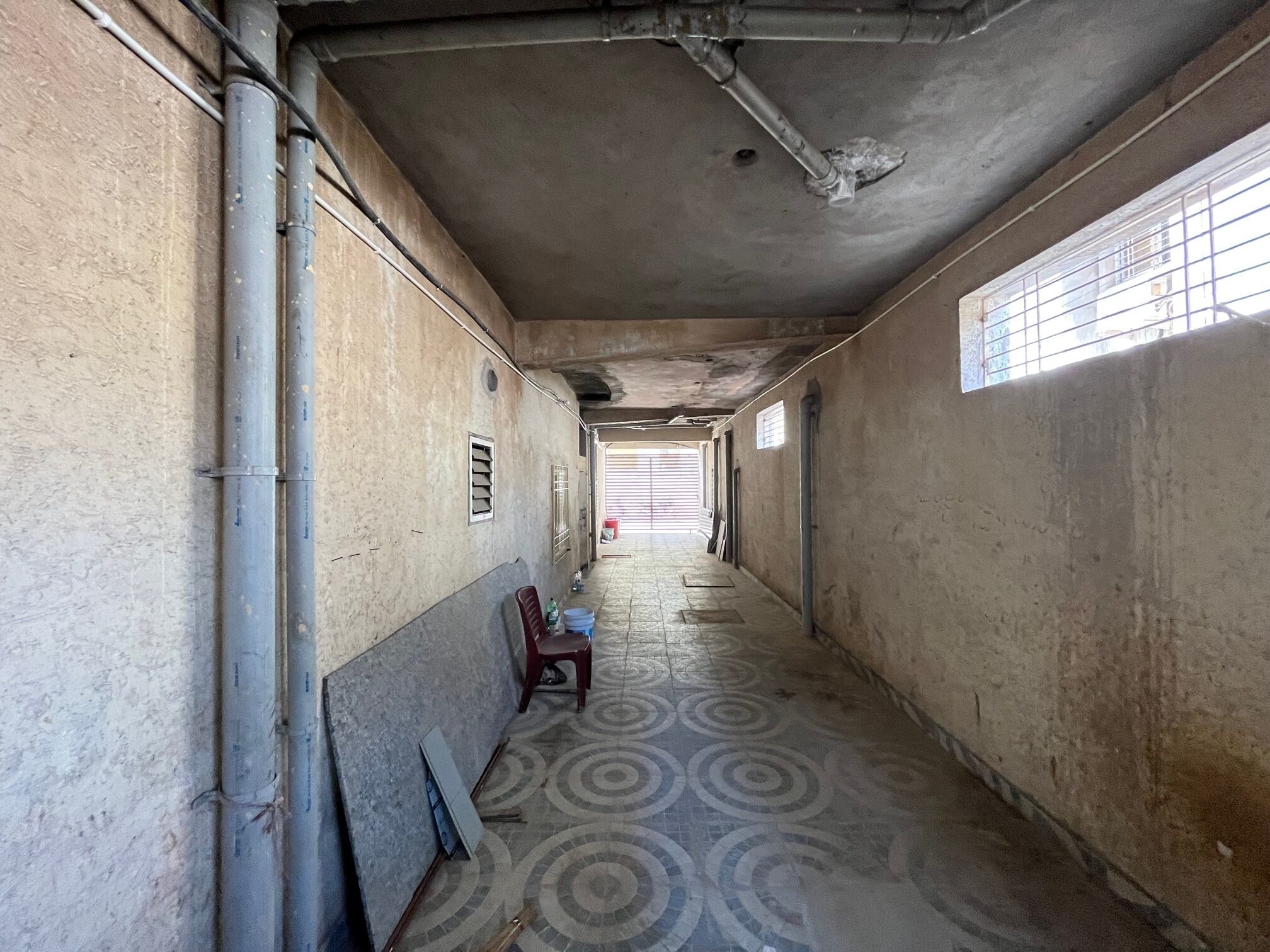 A long, narrow hallway with a patterned floor, concrete walls, and a window on the right side.