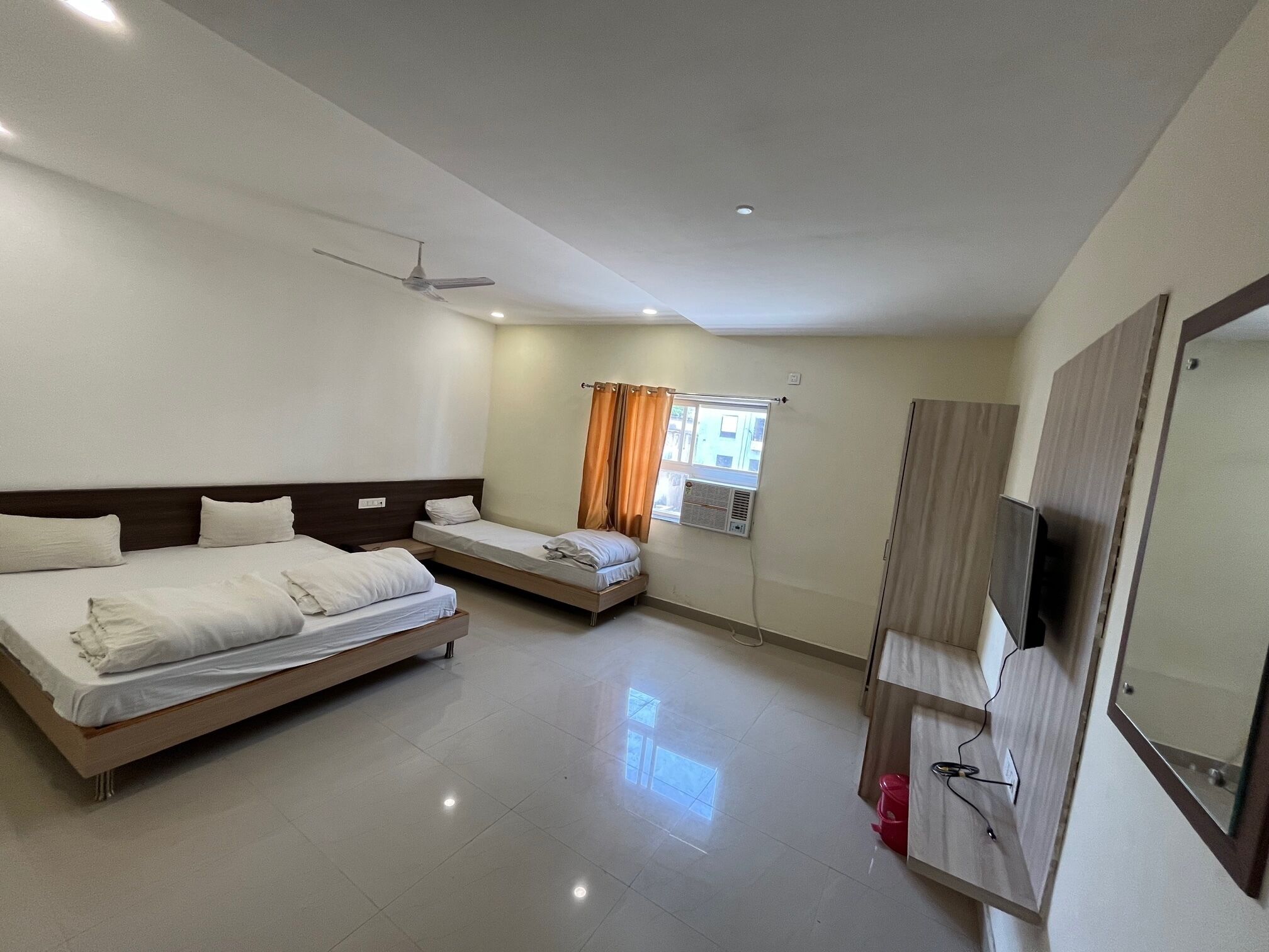 Habitación de hotel con dos camas, un ventilador de techo, una ventana con cortinas, un televisor y un cesto de basura rojo.