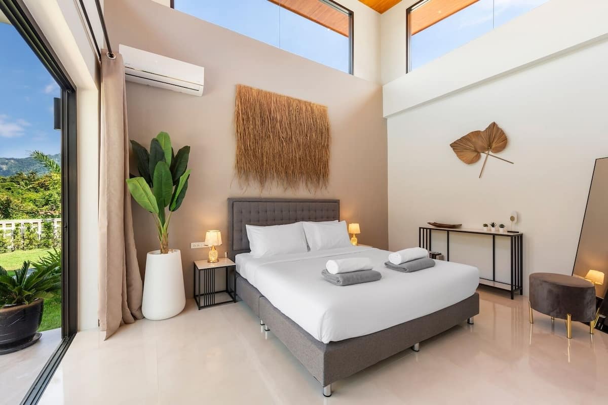 Quarto moderno com uma cama grande, mesas de cabeceira, um vaso com planta e vista para o exterior.