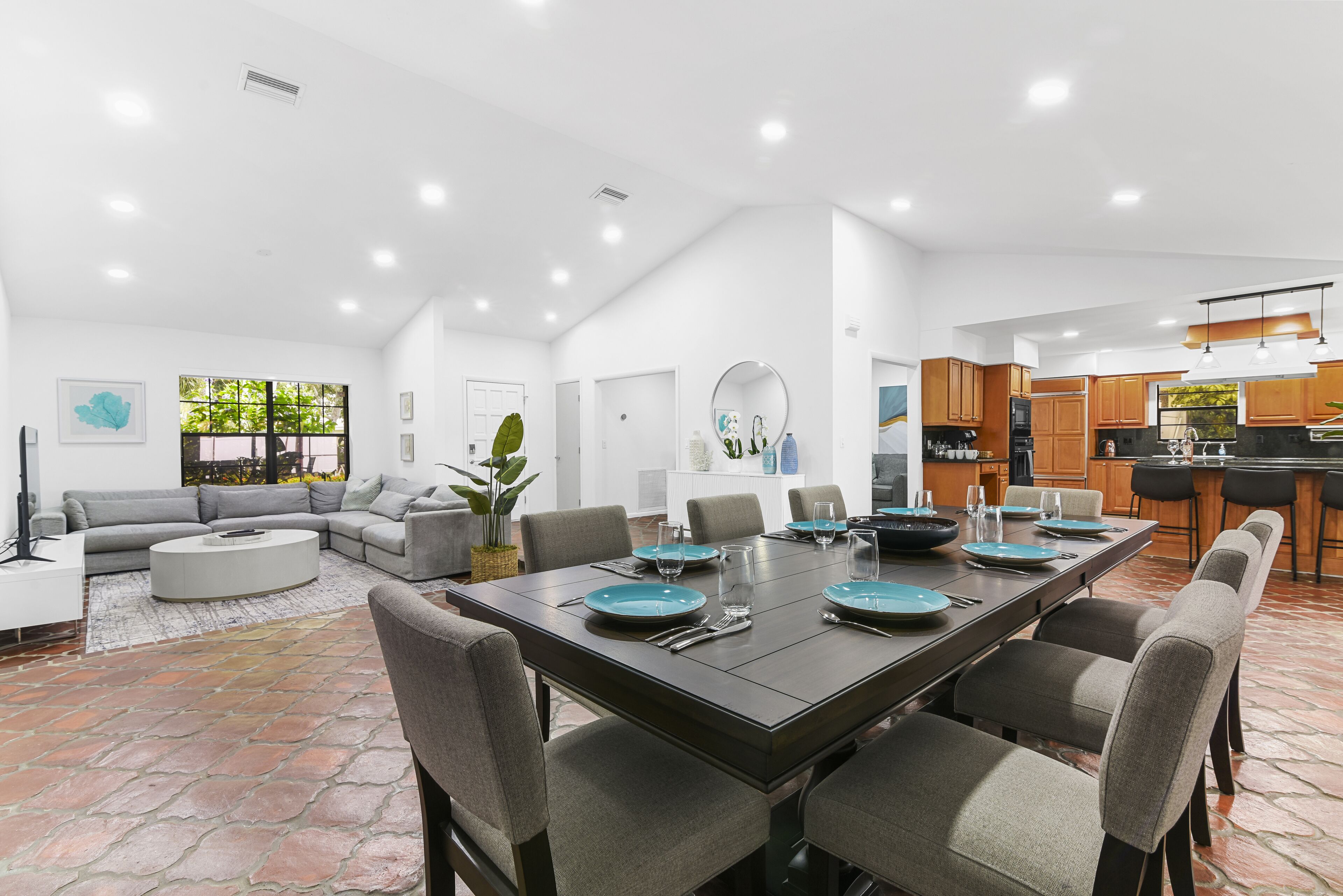 A modern open-plan living and dining area with a large dining table set for six, surrounded by grey upholstered chairs.