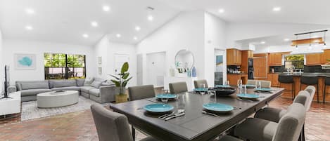 A modern open-plan living and dining area with a large dining table set for six, surrounded by grey upholstered chairs.