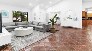 Un soggiorno moderno con un divano ad angolo grigio, un tavolino da caffè bianco e un tappeto decorato. Una grande finestra offre vista su alberi e una porta conduce a un'altra stanza.