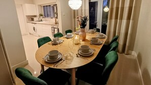 Zona pranzo con tavolo rotondo apparecchiato per sei persone, sedie imbottite verdi, un vaso con fiori e una cucina sullo sfondo.