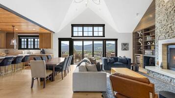 A spacious loft with a high ceiling, a dining area with a long table and chairs, a seating area with sofas and a fireplace, and a view of the mountains through large windows.