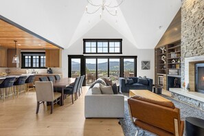 Un loft espacioso con techos altos, un área de comedor con una mesa larga y sillas, una sala de estar con sofás y chimenea, y vistas a las montañas a través de grandes ventanales.